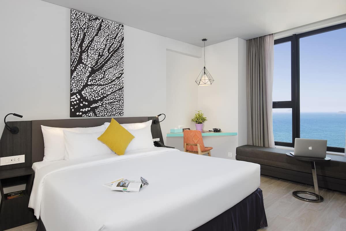 Quarto de hotel moderno com uma cama grande, uma mesa com um laptop, vista para o mar e uma obra de arte decorativa na parede.