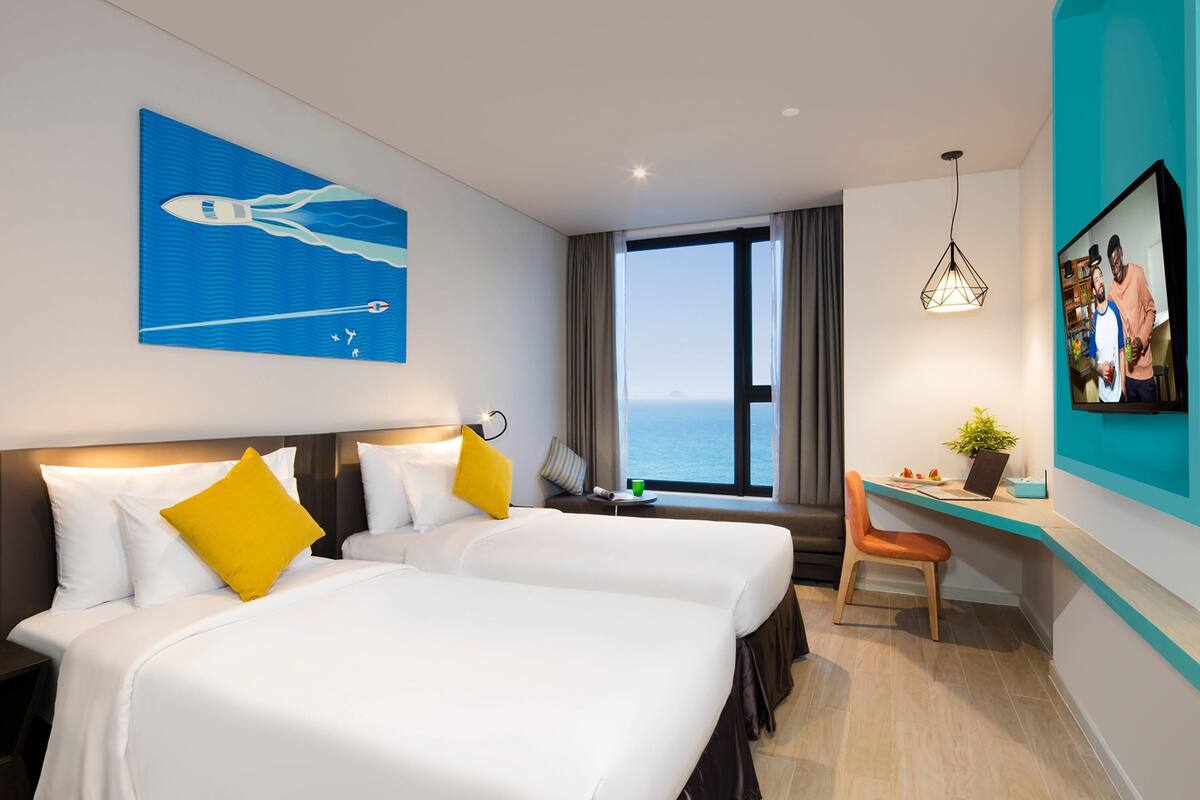 Quarto de hotel com duas camas, uma escrivaninha, televisão e uma janela grande com vista para o mar.
