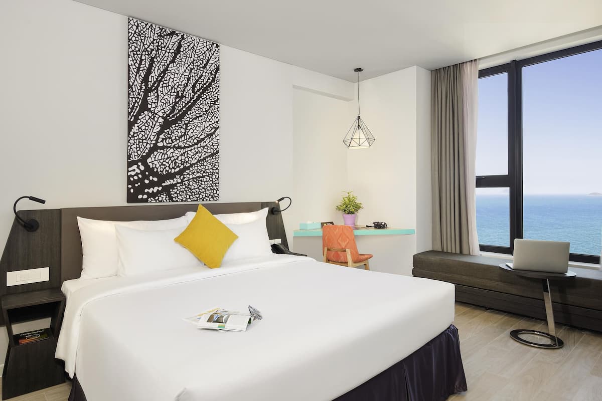A hotel room with a bed, a desk with a laptop, a chair, a potted plant, and a view of the sea.