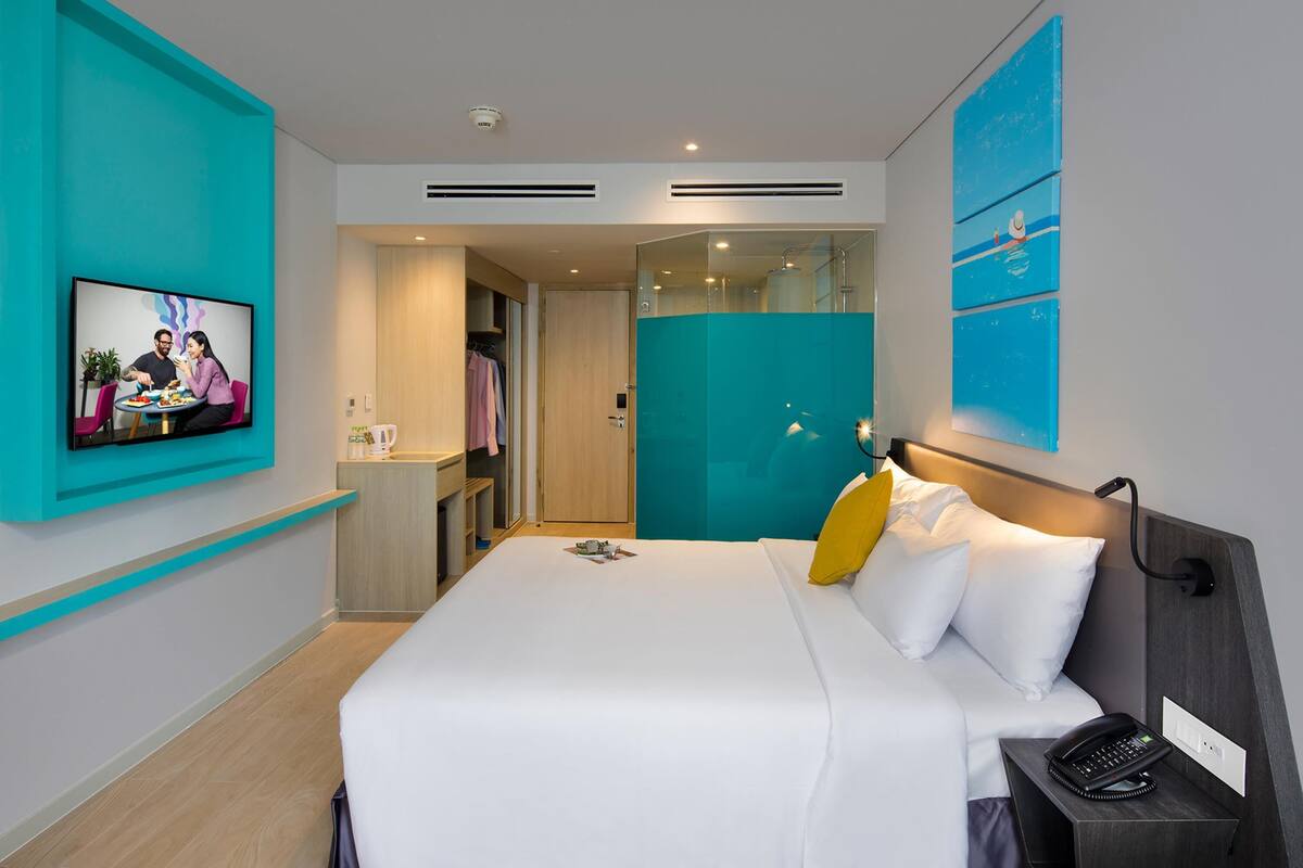 Quarto de hotel moderno com cama, telefone ao lado, TV na parede e banheiro visível através de uma porta aberta.