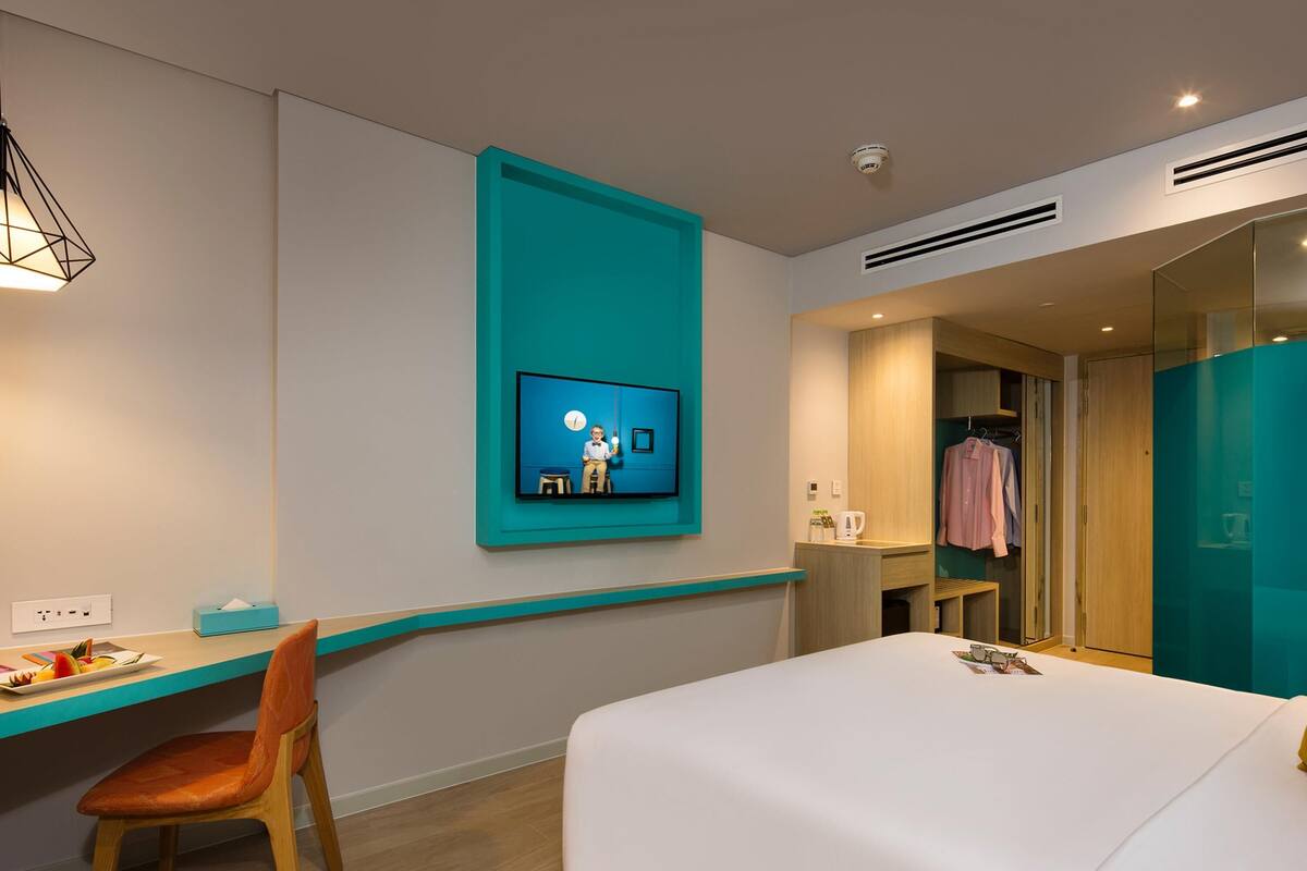 Quarto de hotel com cama, escrivaninha, cadeira, televisão na parede e guarda-roupa.