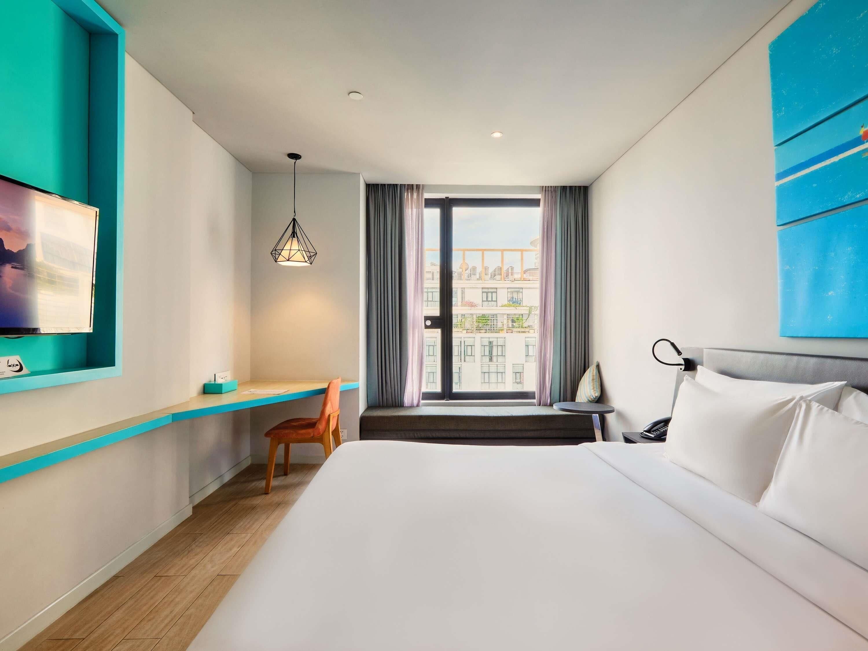 Habitación de hotel con cama, escritorio, silla, televisor y ventana con cortinas.