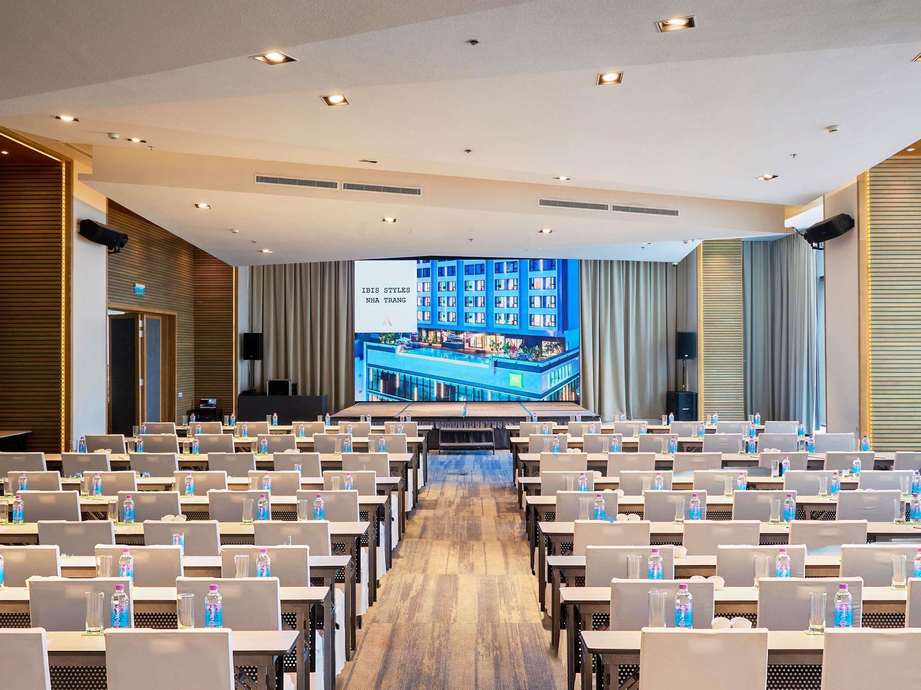 Un salón de conferencias con filas de mesas y sillas, una pantalla grande que muestra una imagen de un edificio y un escenario con un atril.