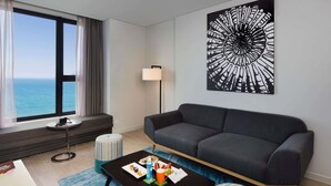 A modern living room with a large window, a black coffee table, a grey sofa, a striped ottoman, and a wall-mounted artwork. - ibis Styles Nha Trang (Nha Trang)