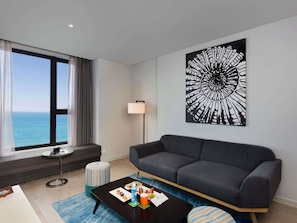 A modern living room with a large window, a black coffee table, a grey sofa, a striped ottoman, and a wall-mounted artwork. - ibis Styles Nha Trang (Nha Trang)
