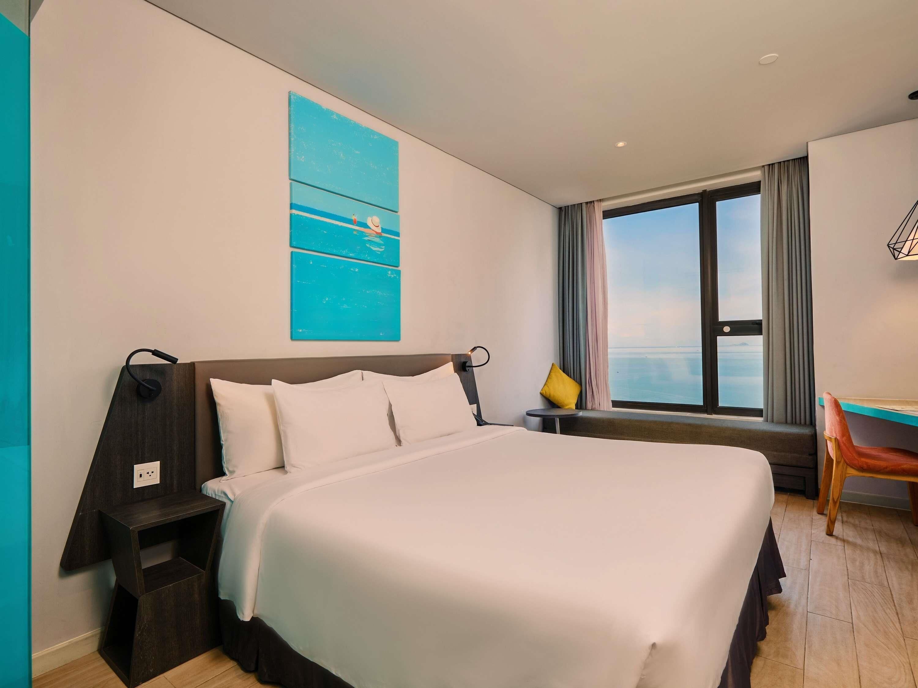 Ein Hotelzimmer mit einem großen Bett, einem Schreibtisch, einem Stuhl, einem Fenster mit Blick und einem blauen Kunstwerk an der Wand.
