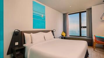 Ein Hotelzimmer mit einem großen Bett, einem Schreibtisch, einem Stuhl, einem Fenster mit Blick und einem blauen Kunstwerk an der Wand.