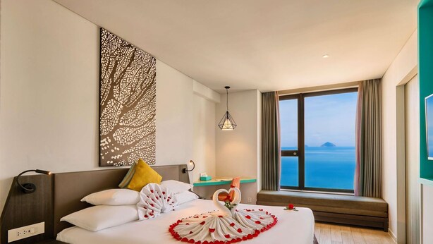 A hotel room with a bed, a nightstand, a wall art piece, a window with a view, and a small table with a lamp. - ibis Styles Nha Trang (Nha Trang)