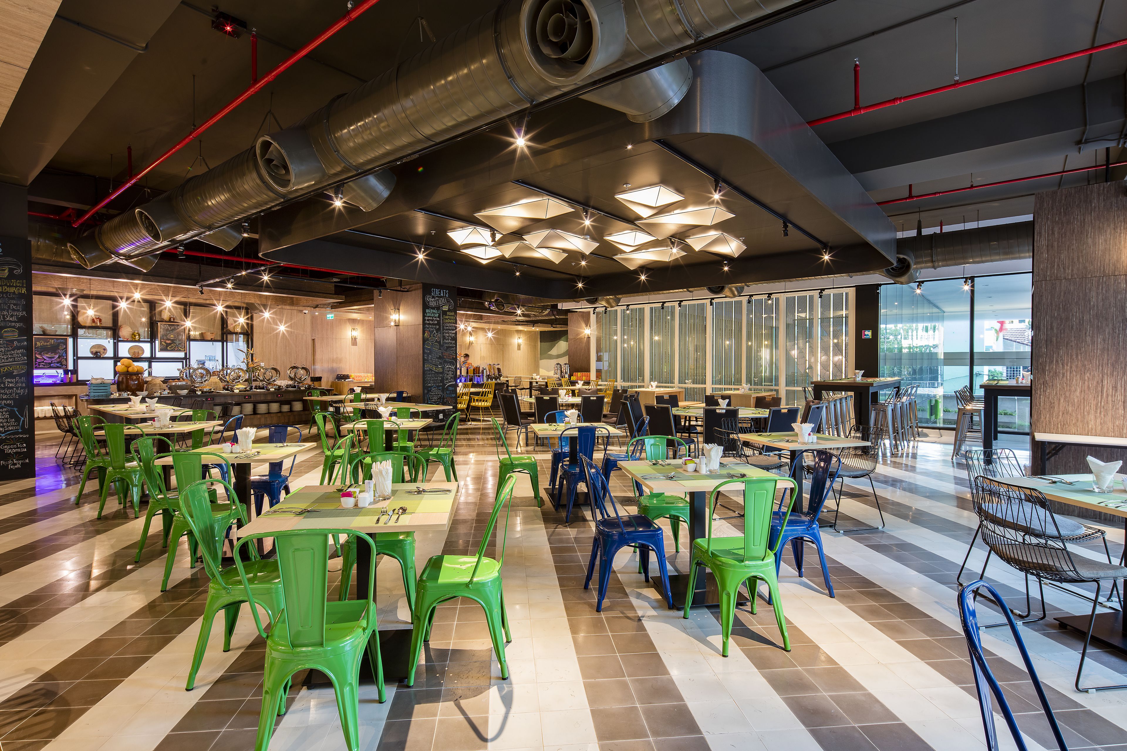 A spacious cafeteria with high ceilings, exposed ductwork, and modern lighting fixtures. Tables and chairs are arranged in an open layout, with a view of the city through large windows.