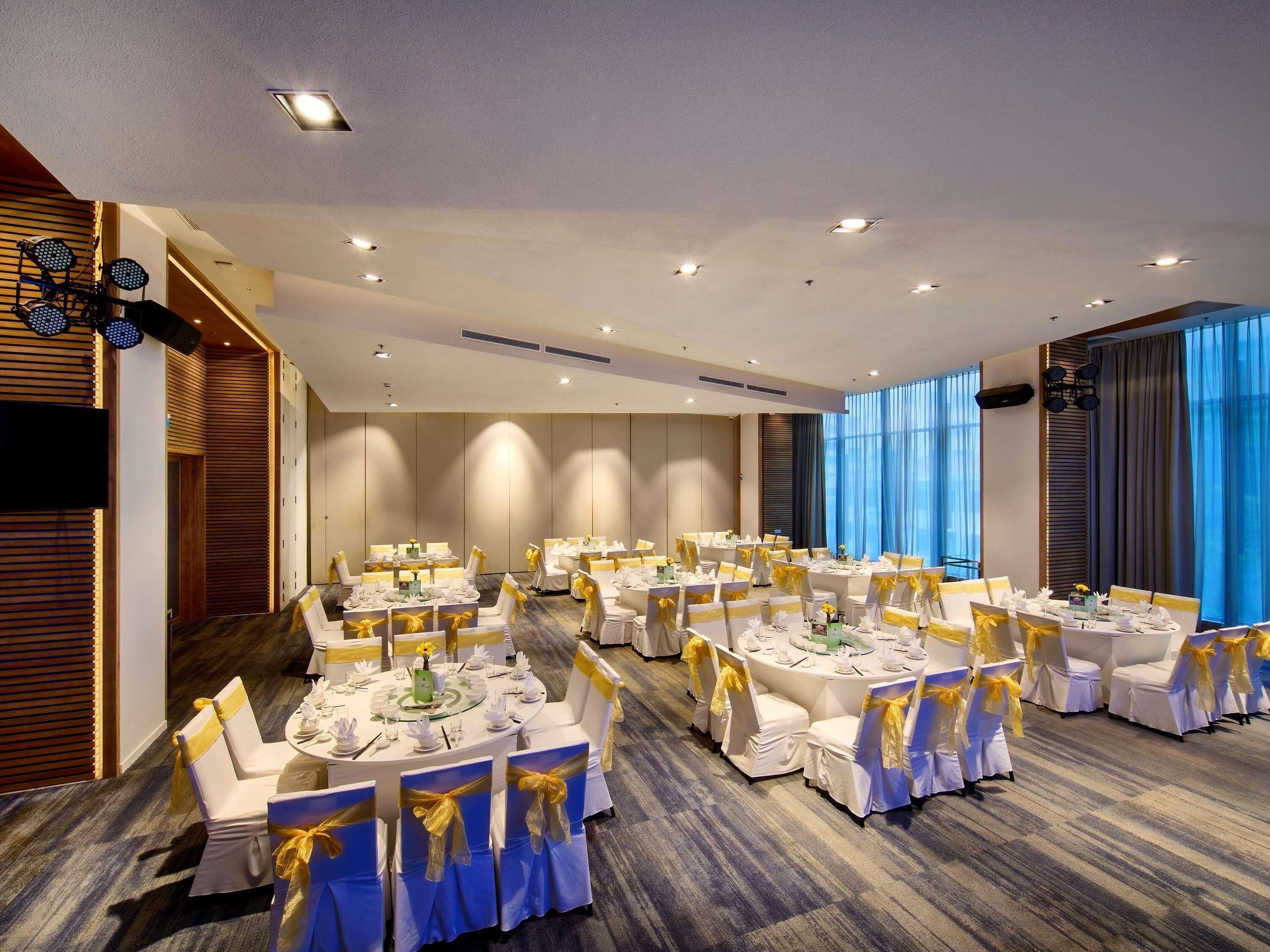 Uma ampla sala de banquetes com mesas redondas, cadeiras brancas com fitas amarelas e uma grande janela com cortinas azuis.