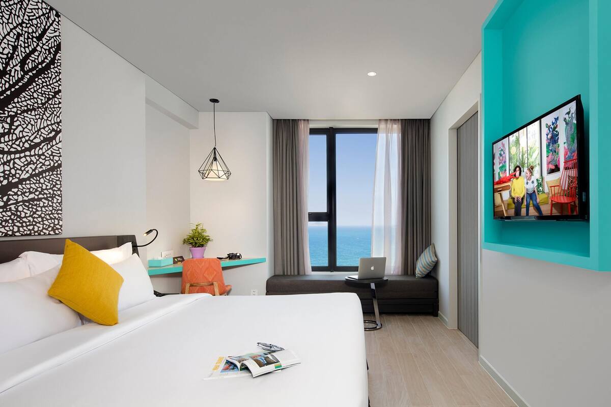 Quarto de hotel moderno com uma cama grande, uma mesa com um laptop, vista para o oceano e um quadro na parede.