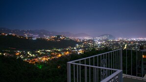 Vistes nocturnes d'una ciutat amb una serralada de muntanyes al lluny, un balcó amb barana i llums de la ciutat al fons.