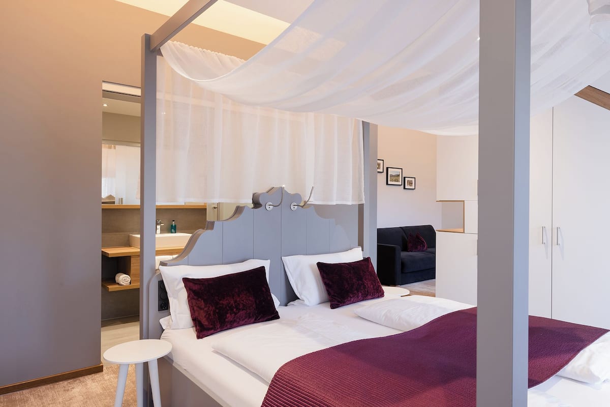 A modern hotel room with a bed, a small table, a wardrobe, and a bathroom visible through a glass partition.