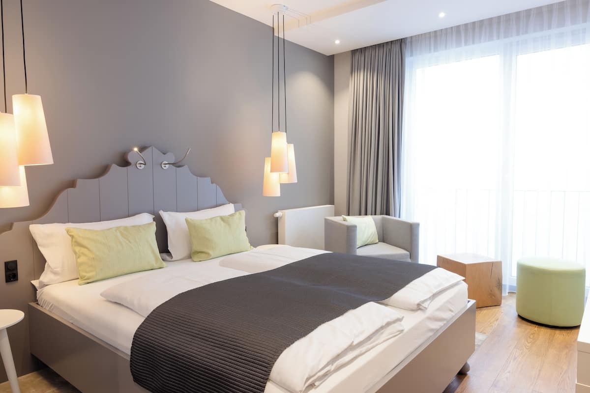 A modern hotel room with a large bed, a chair, a small ottoman, and a view of the outdoors through the window.