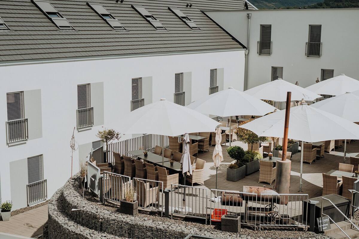 An outdoor seating area with tables and umbrellas, surrounded by a stone wall and a building with multiple windows.