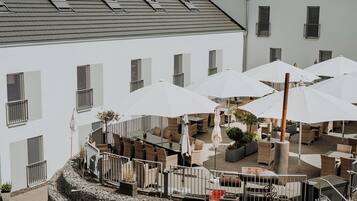 An outdoor seating area with tables and umbrellas, surrounded by a stone wall and a building with multiple windows.