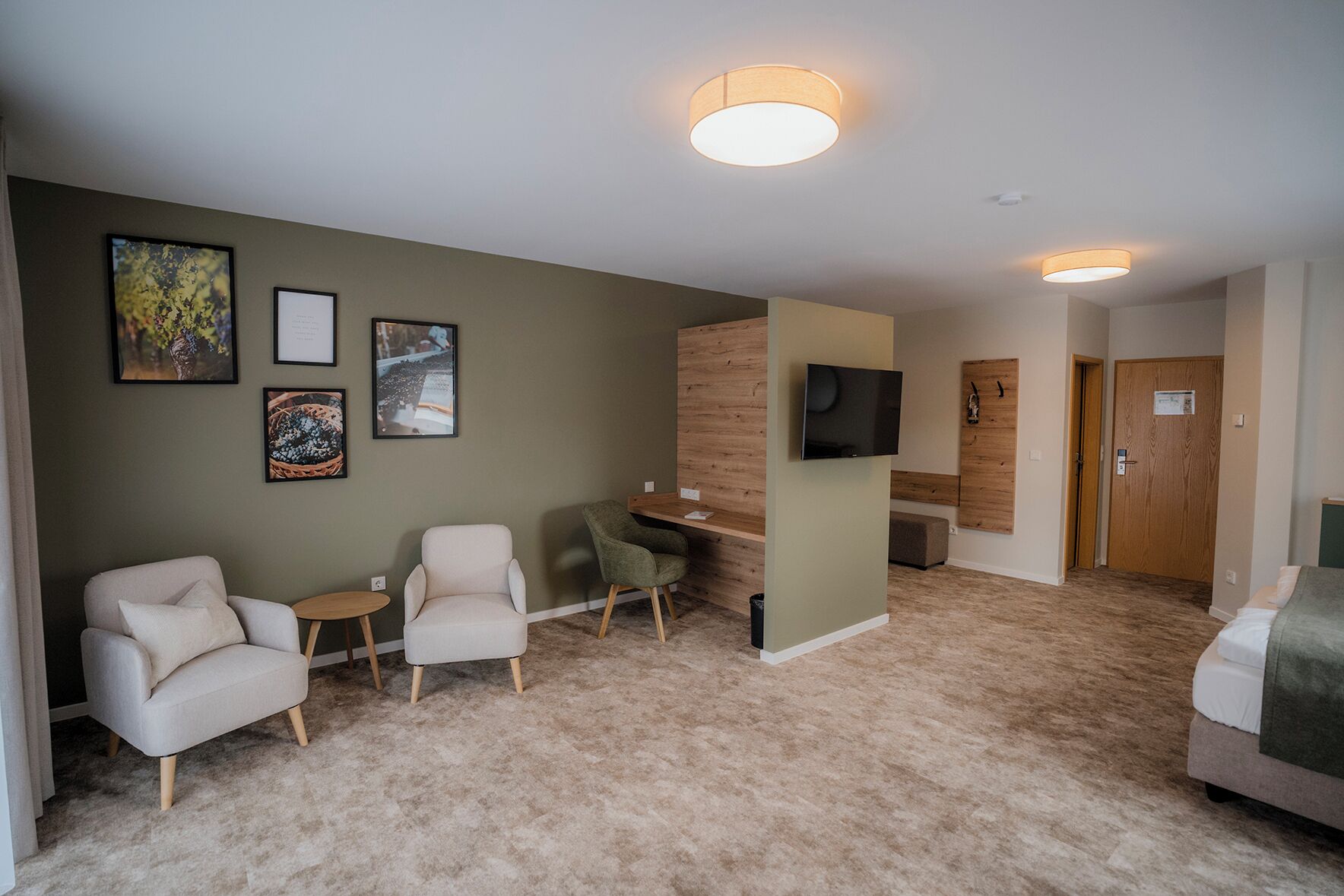 Habitación de hotel con piso alfombrado, dos sillones, una mesita y un televisor fijado a la pared, además de una puerta de madera.