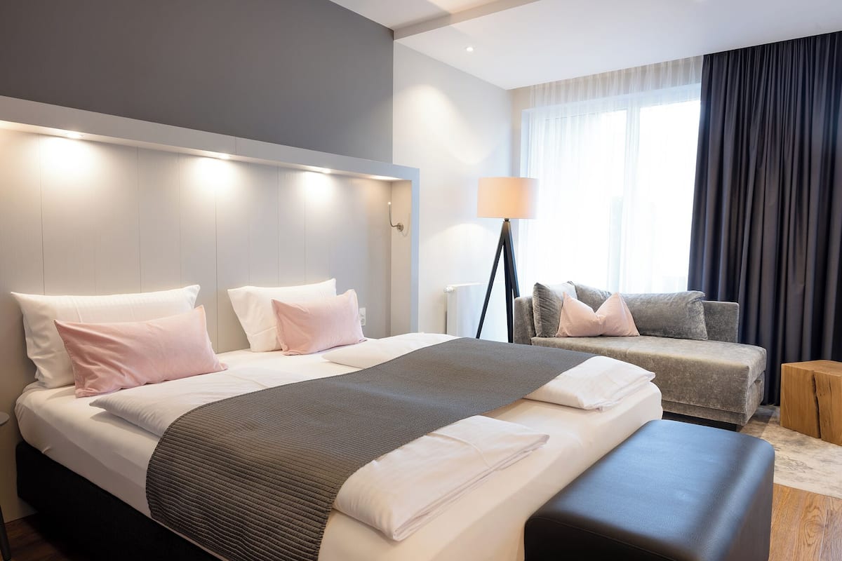 A modern hotel room with a large bed, a sofa, a floor lamp, and a view of the city through the window.