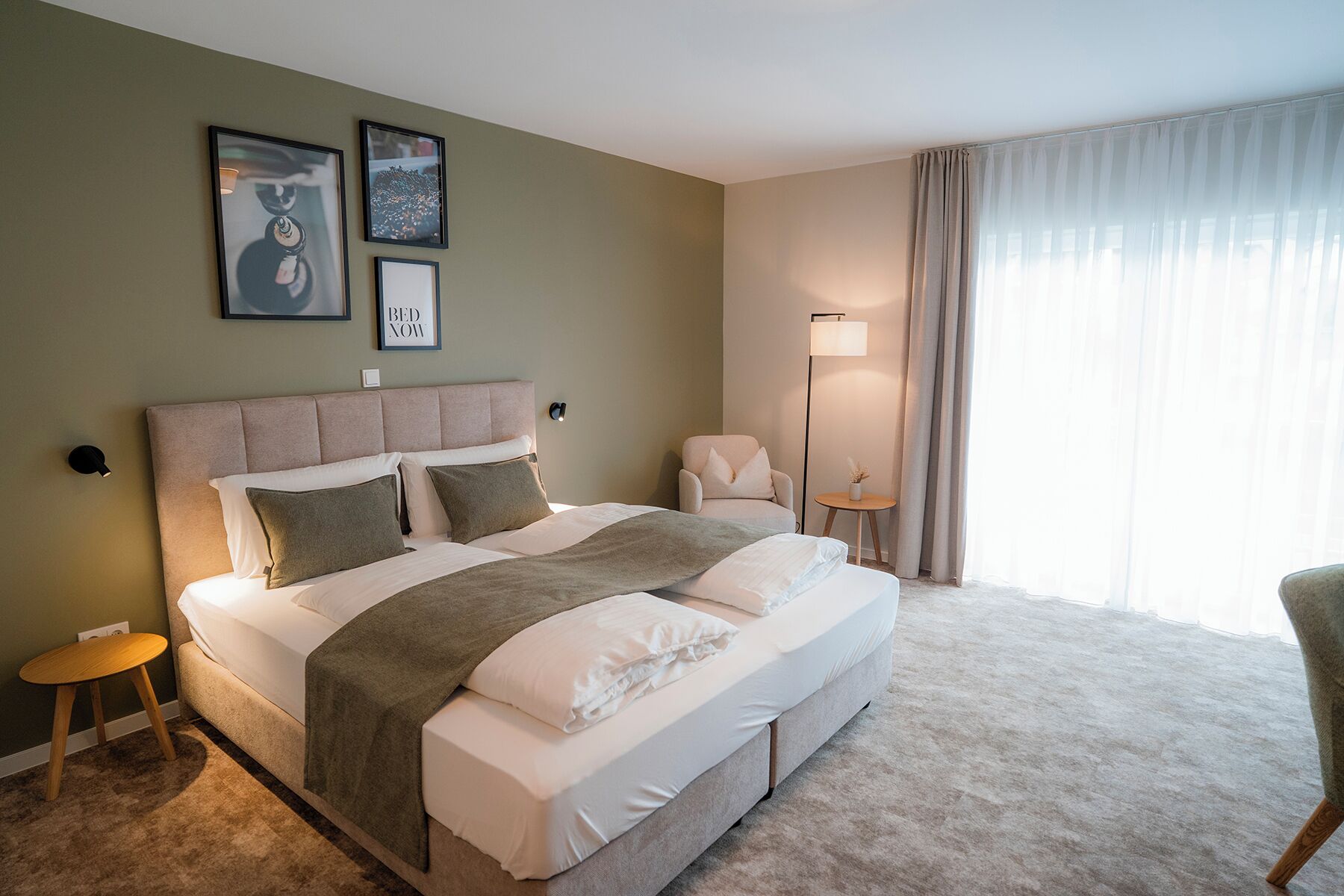 A hotel room with a large bed, a bedside table, a chair, a floor lamp, and a window with curtains.