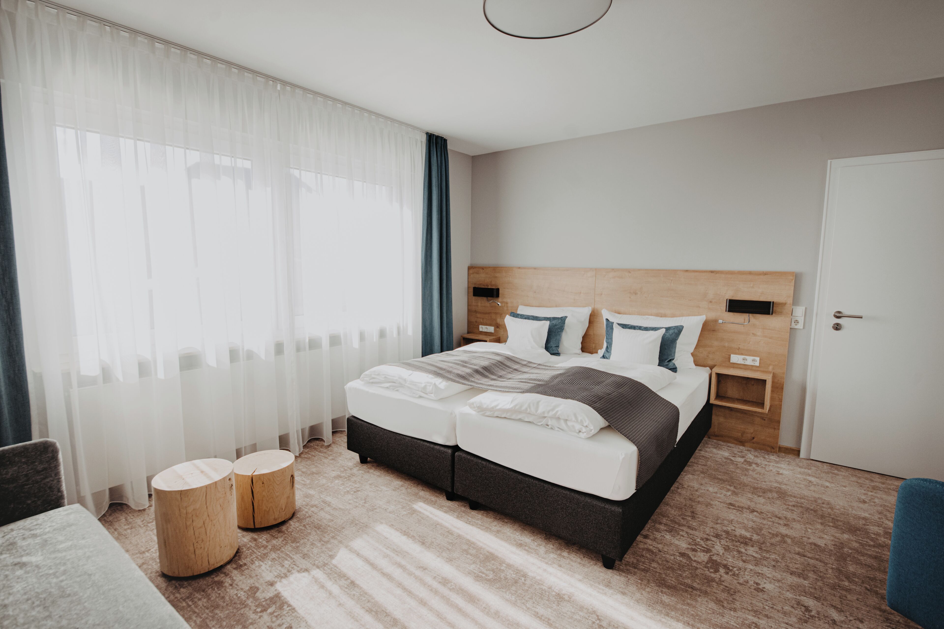 Quarto de hotel moderno com uma cama grande, mesas de cabeceira, encosto de madeira e vista para o exterior através de cortinas translúcidas.