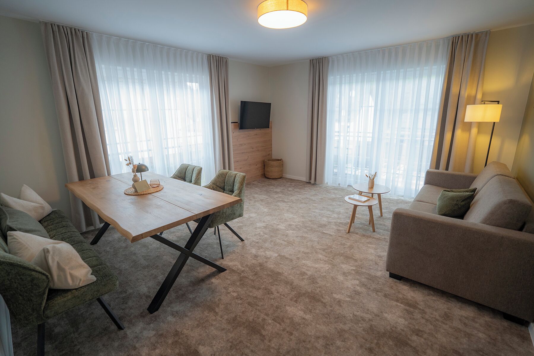 A modern living room with a wooden dining table, green upholstered chairs, a beige sofa, and a small side table with a lamp.