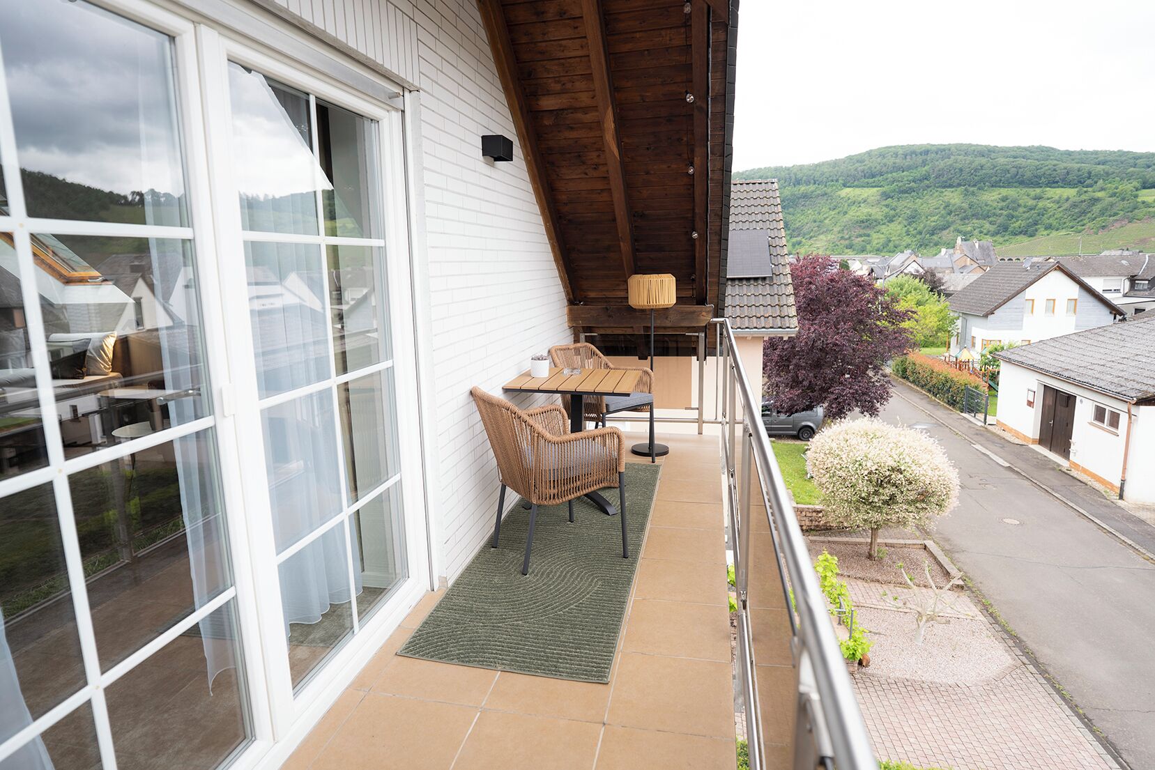 A balcony with a table and chairs, a small rug, and a view of a residential street and hills.