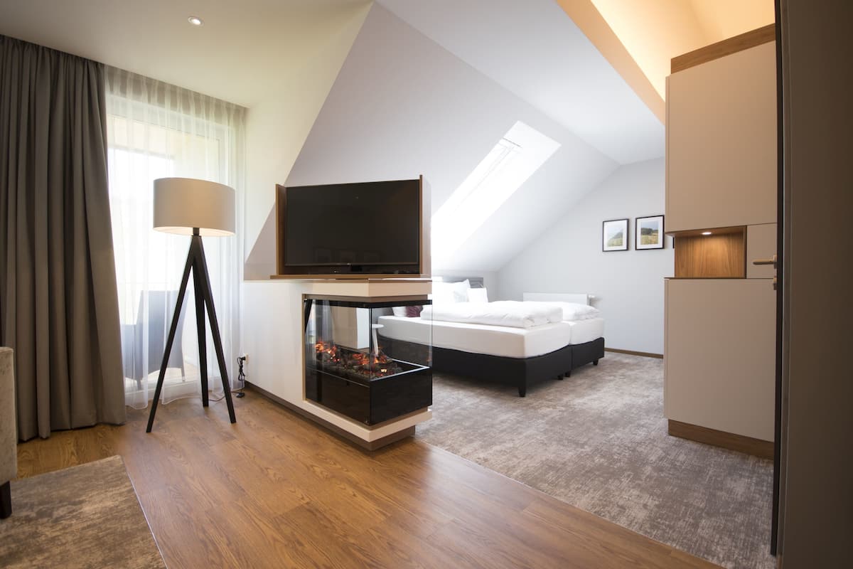 A modern hotel room with a large bed, a flat-screen TV, a fireplace, and a large window with curtains.