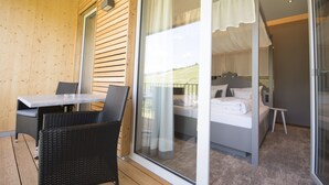 A room with a bed, a small table, and a chair, with a view of a green landscape through a glass door.