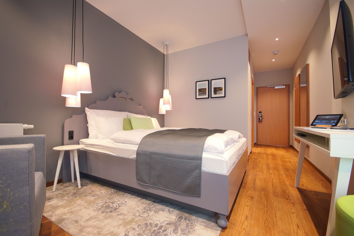 Ein modernes Hotelzimmer mit Bett, Nachttisch, Sessel und Schreibtisch mit Computer.