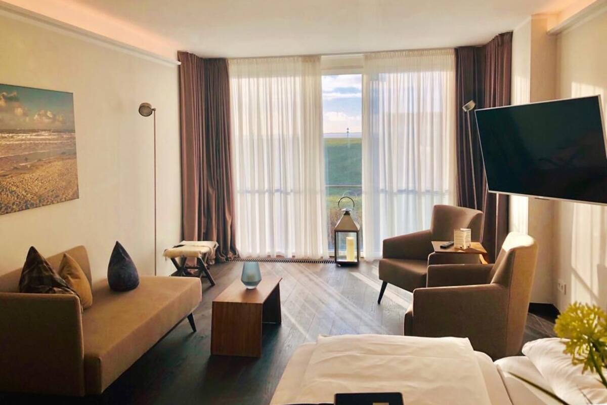 Habitación de hotel con cama, sofá, dos sillones, televisión y un amplio ventanal con cortinas.