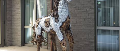 A sculpture of a horse made from natural materials, positioned on a tiled floor in front of a building with large windows and a brick facade.