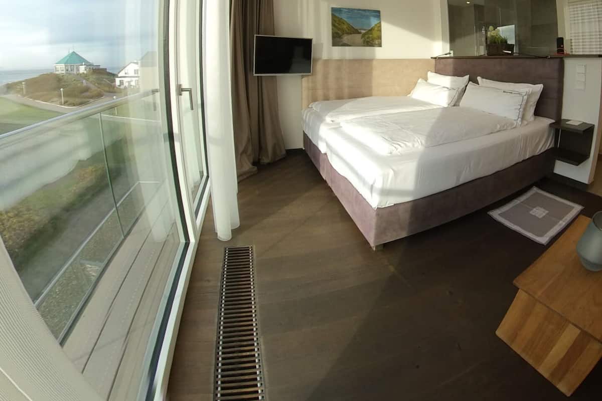 Una habitación de hotel moderna con una cama grande, un televisor de pantalla plana, una mesita de noche y vistas al mar a través de un ventanal amplio.