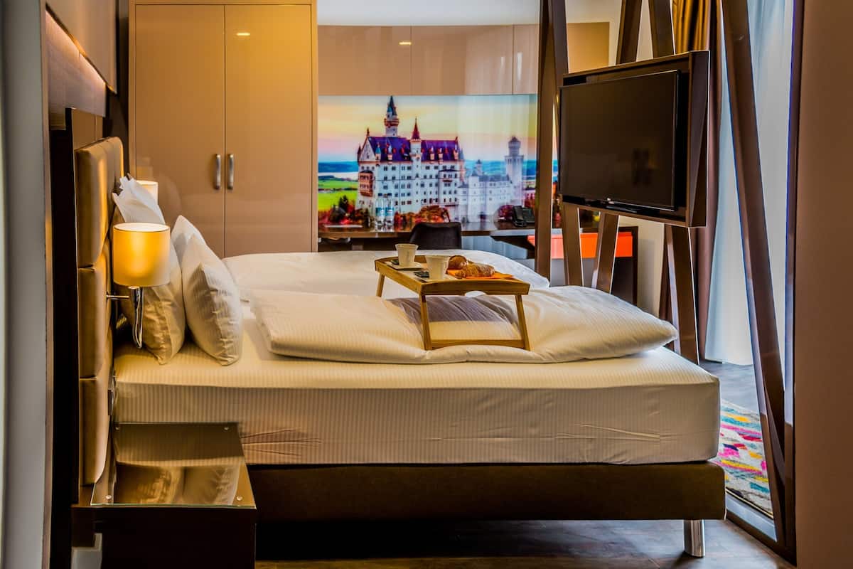 A modern hotel room with a large bed, a bedside table with a tray of breakfast, a TV, and a scenic view of a castle through the window.
