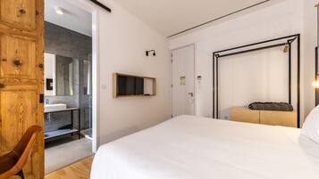 A modern hotel room with a large bed, a wooden wardrobe, a bathroom with a glass shower, and a mounted television.