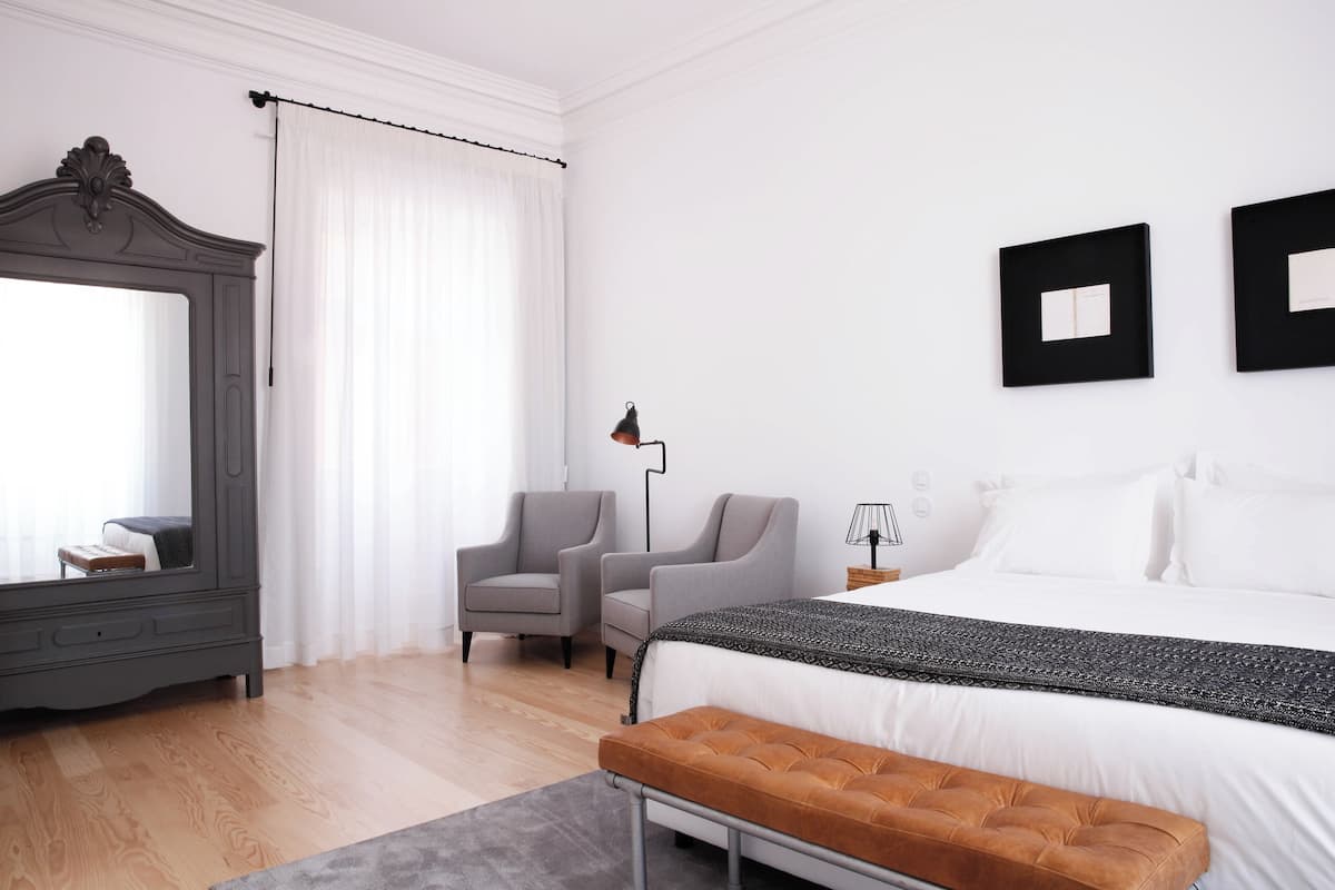 Une chambre d’hôtel moderne, dotée d’un grand lit, de deux fauteuils, d’un banc, d’un miroir et d’une table de chevet avec une lampe.