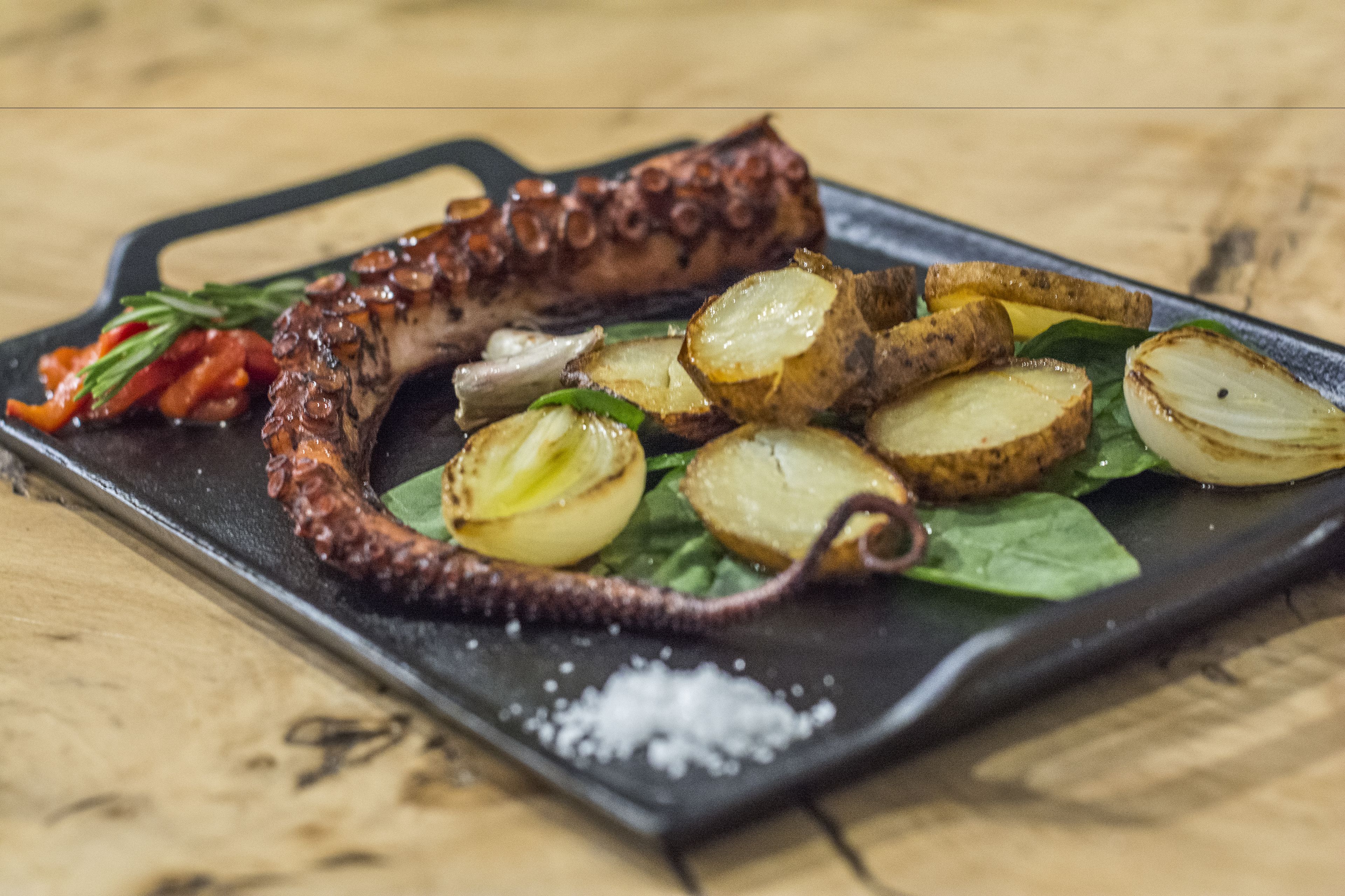 A dish of grilled octopus with potatoes and greens on a black plate.