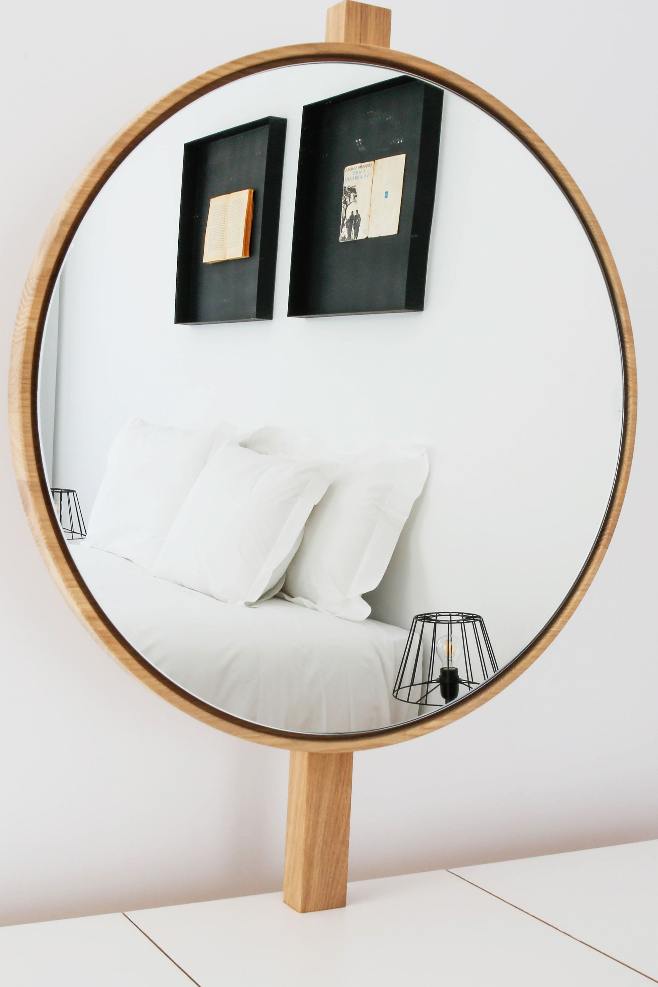 Un miroir rond, doté d’un cadre en bois et posé sur un support, reflète un lit avec une literie blanche et une lampe.