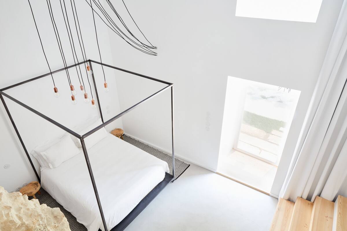 Une chambre à coucher moderne avec un cadre de lit en verre, une literie blanche et une vue sur l’extérieur grâce à une grande fenêtre.