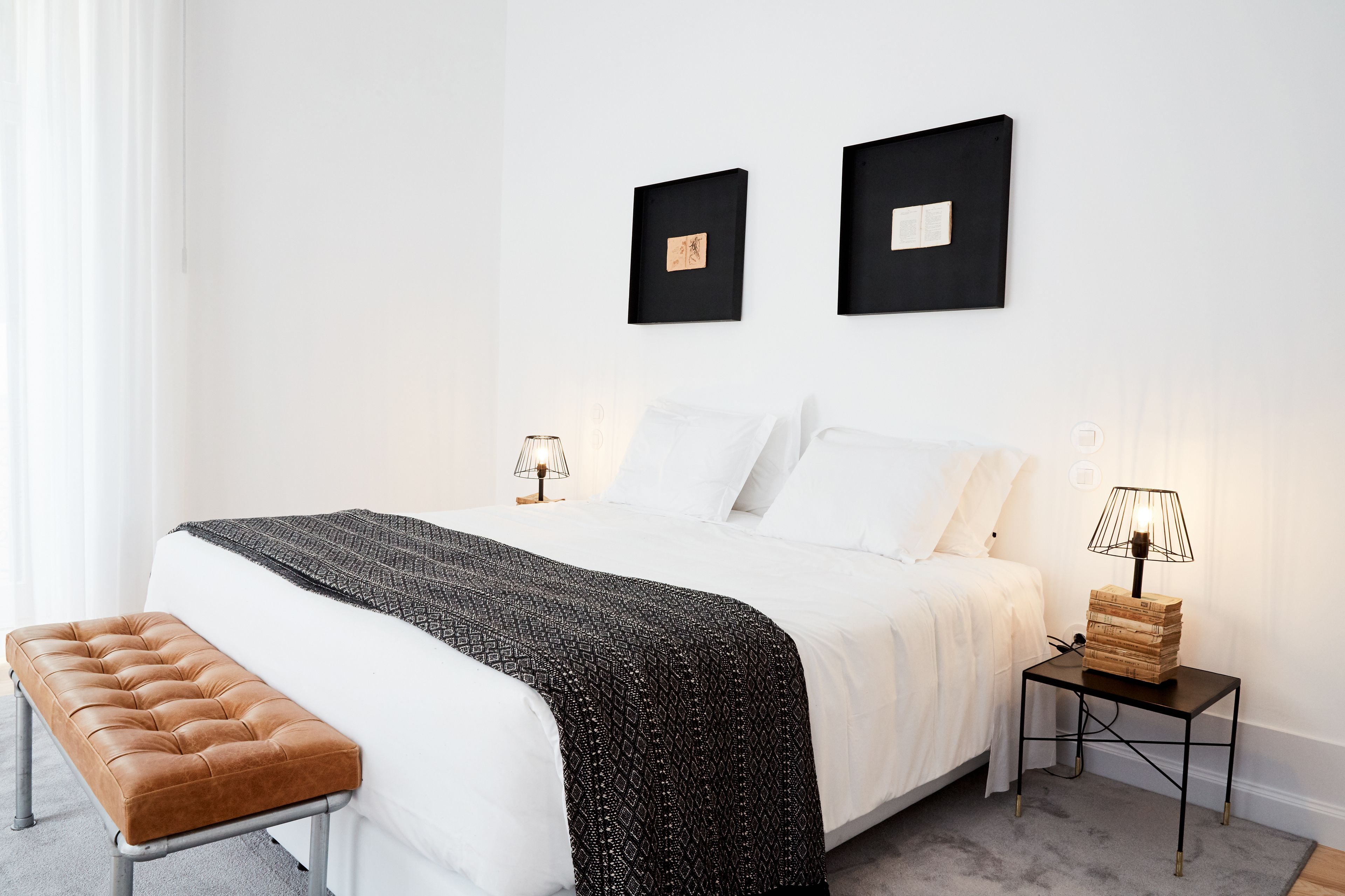 A neatly arranged bedroom with a bed, a bench, a bedside table with a lamp, and two framed pictures on the wall.