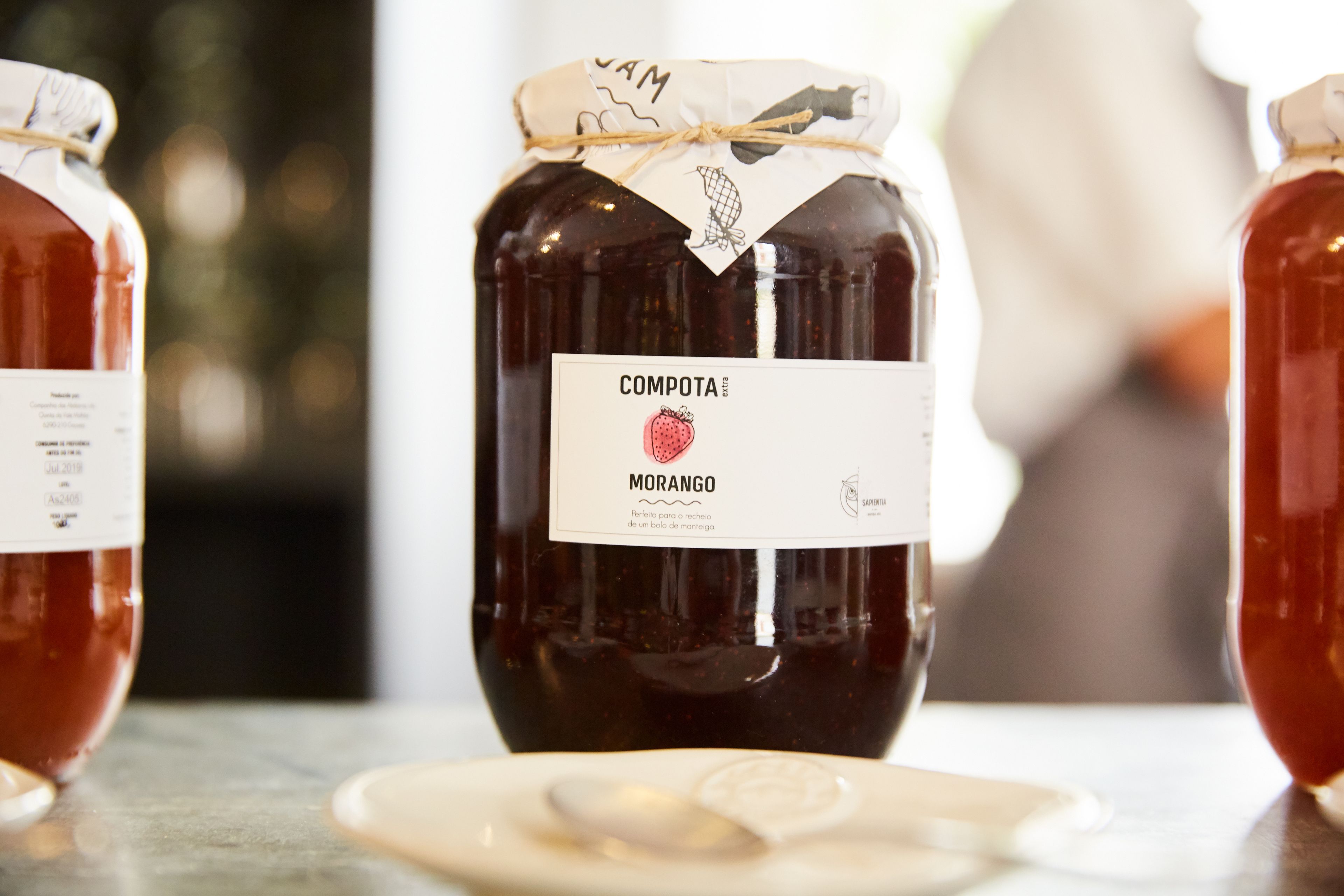 A jar of compota morango with a label, placed on a table with a spoon and a plate nearby.