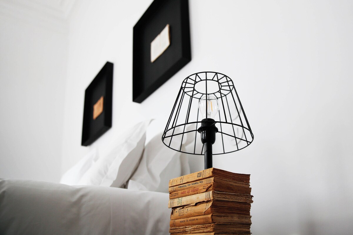 Een bed met witte lakens, een zwarte metalen lamp met een heldere kap en een stapel boeken op een nachtkastje.