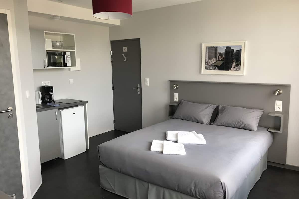 A modern hotel room with a grey bed, a small kitchenette, a dark door, and a framed cityscape picture on the wall.