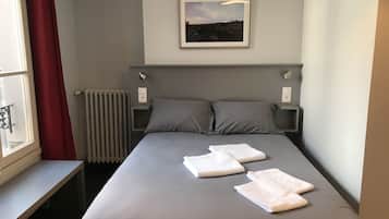 A neatly made bed with pillows and two folded towels, a framed picture on the wall, a window with a red curtain, and a radiator.