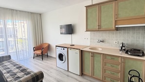A compact living space with a sofa, a washing machine, a small kitchen with cabinets, a television, and a window with curtains. - Fora Apart Otel (Cesme)