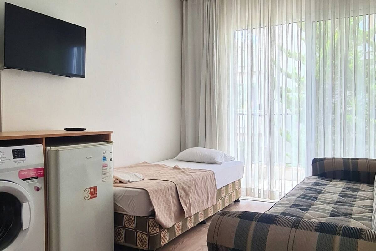 Una habitación con cama, sofá, lavadora, refrigerador y televisor de pantalla plana.