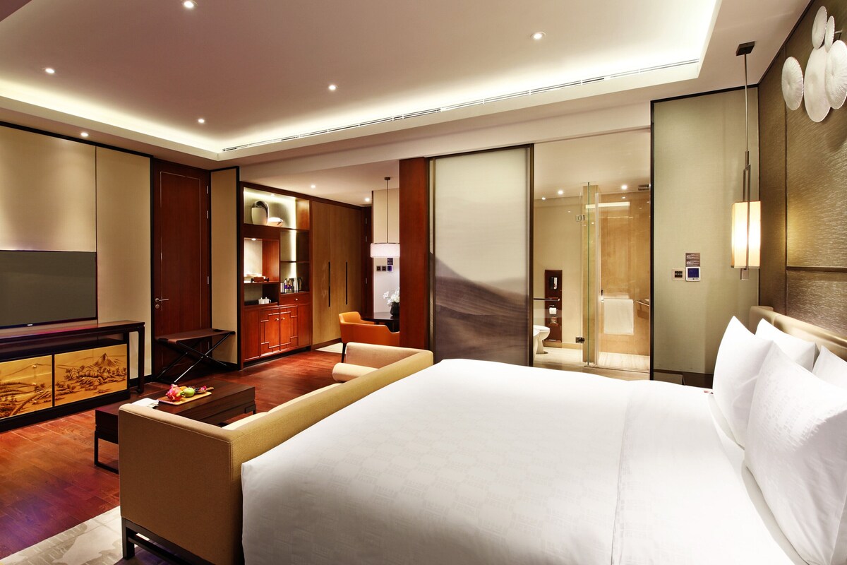 A hotel room with a large bed, a television, a desk with a chair, and a bathroom visible through a glass door.