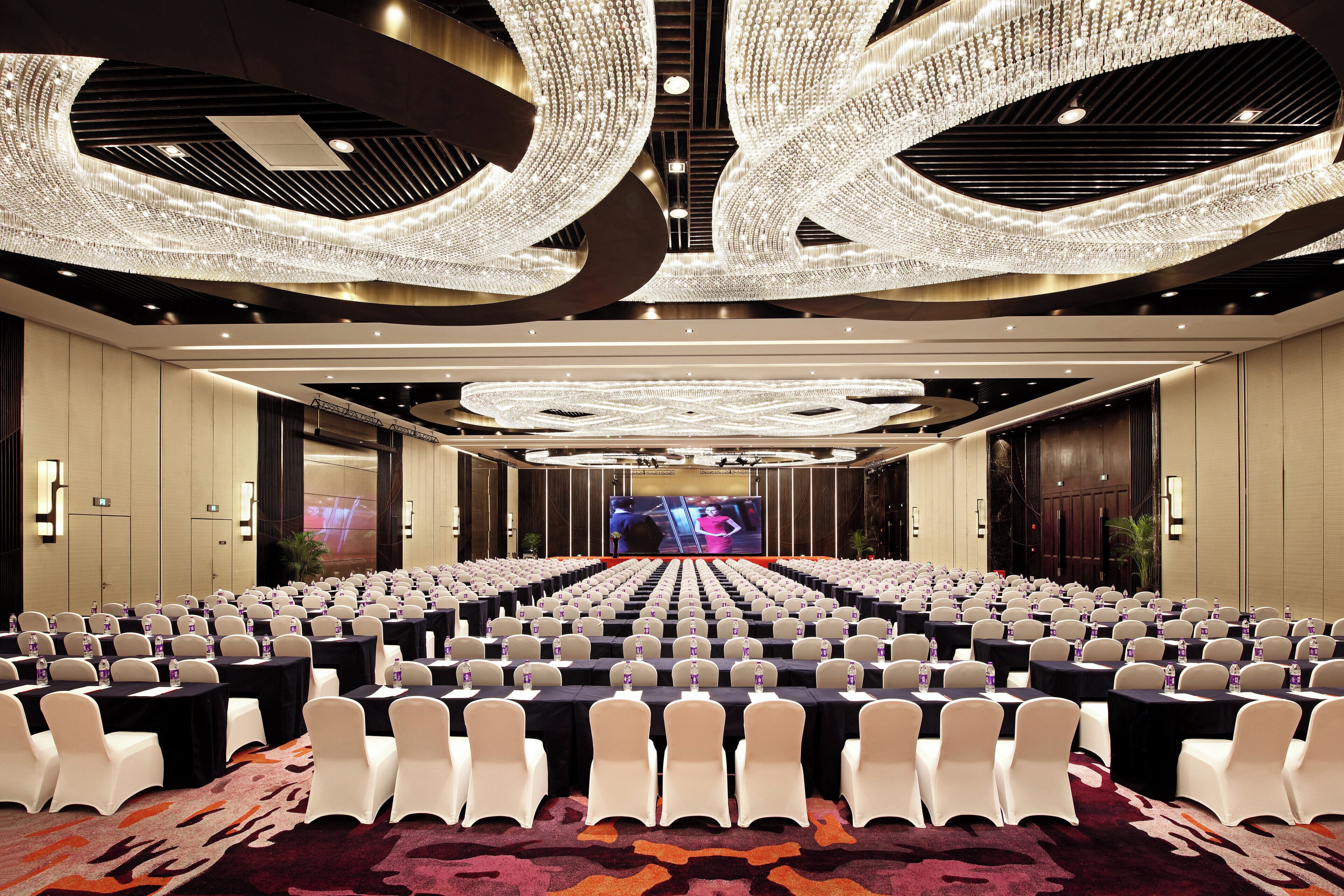 A conference room with rows of tables and chairs, a large screen at the front, and a ceiling with a unique light fixture.