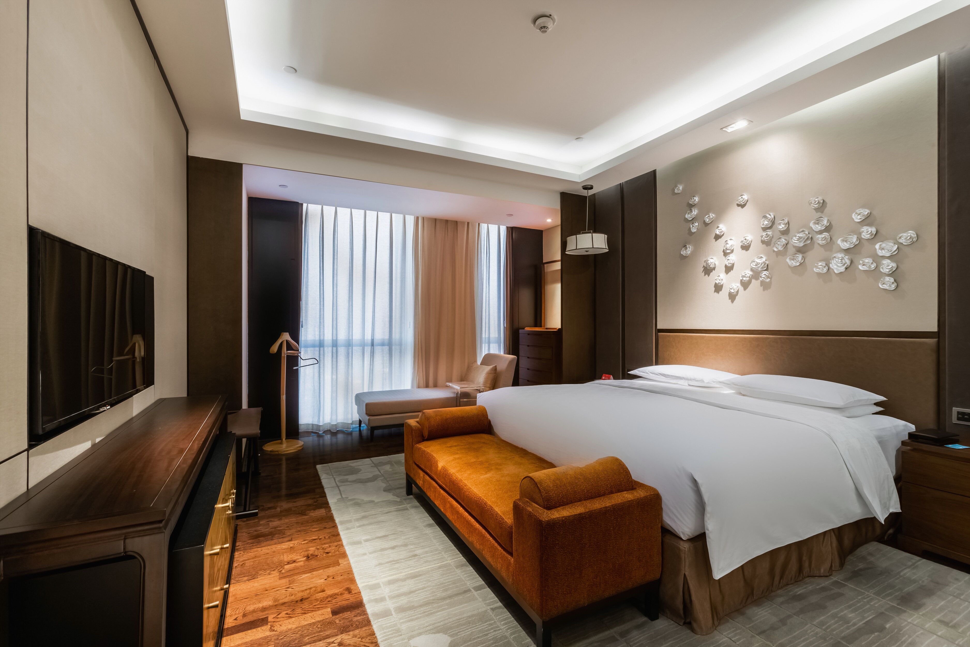 A modern hotel room with a large bed, a sofa, a TV, and a view of the city through large windows.
