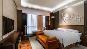 A modern hotel room with a large bed, a sofa, a TV, and a view of the city through large windows.