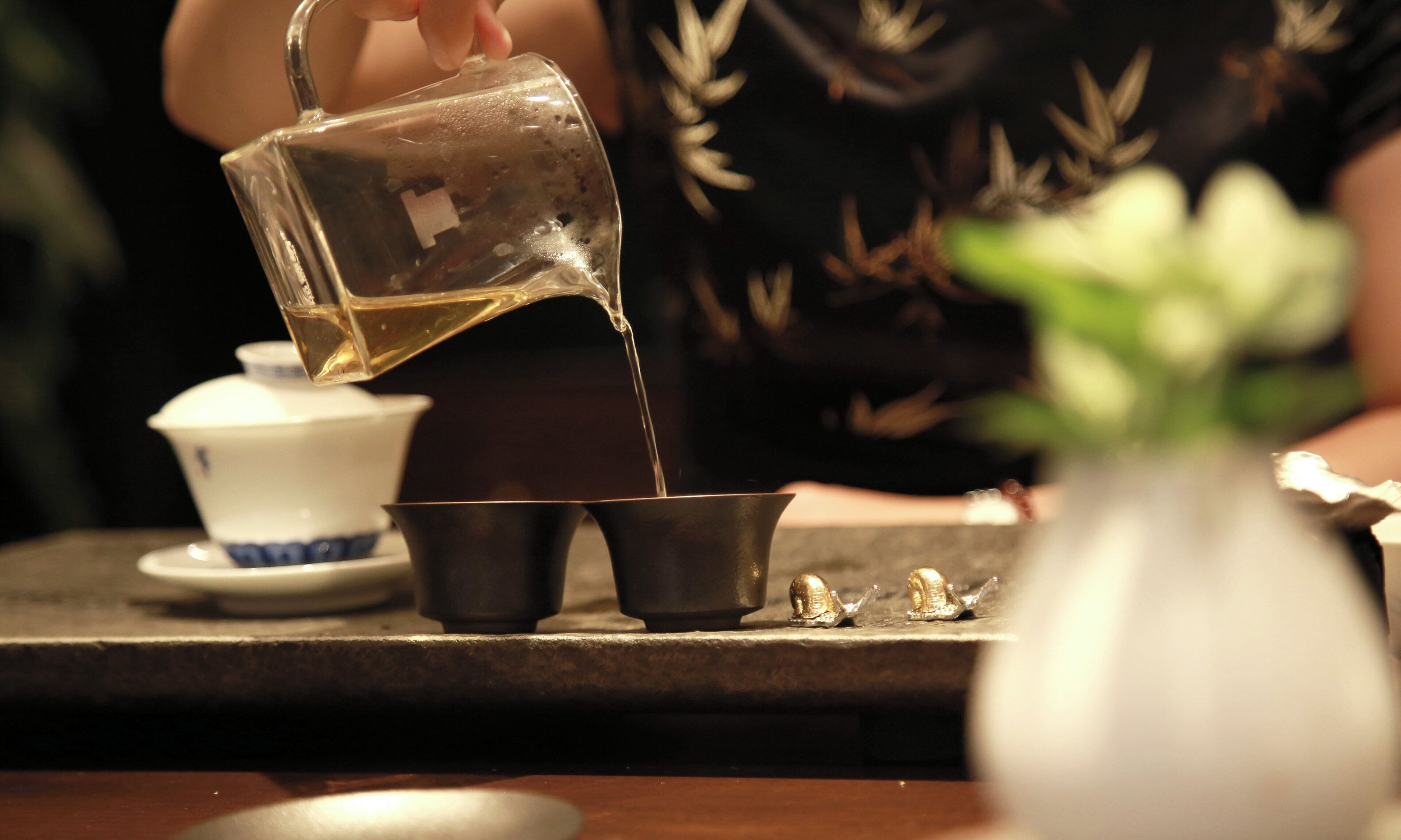 A person pouring a liquid from a jug into a cup, with a teapot and a small bowl on the table.